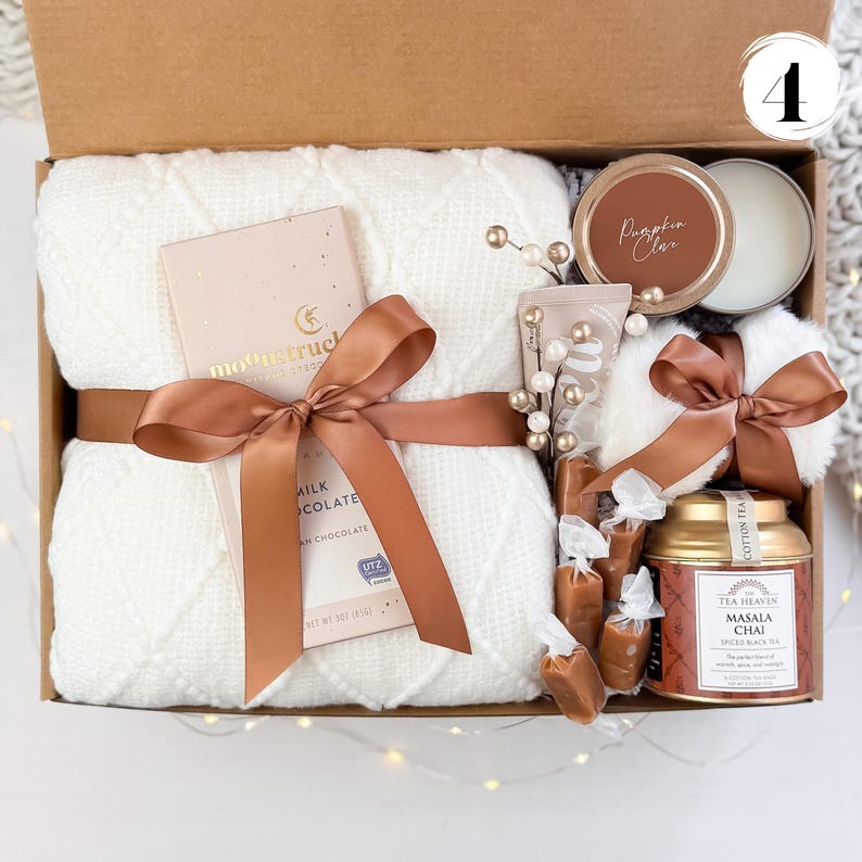 May include: A gift box filled with a white blanket, chocolate bar, candle, tea, and other treats. The box is tied with a brown ribbon. The candle is labeled "Pumpkin Clar".