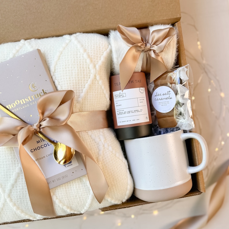 May include: A gift box filled with a cream-colored waffle-knit blanket, a mug with a beige base, a bar of milk chocolate, a gold spoon, tea, and sea salt caramels. The items are tied with beige satin ribbons.