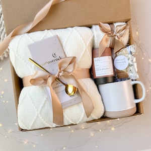 May include: A gift box filled with a cream-colored waffle-knit blanket, a bar of Moonstruck milk chocolate, a gold spoon, a mug, tea, and sea salt caramels. The items are tied with a satin ribbon. The box is brown.