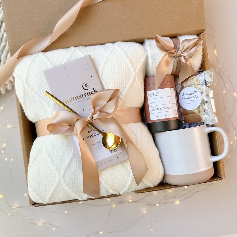 May include: A gift box filled with a cream-colored waffle-knit blanket, a bar of Moonstruck milk chocolate, a gold spoon, a mug, tea, and sea salt caramels. The items are tied with a satin ribbon. The box is brown.