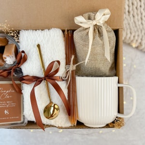 May include: A gift box containing a selection of comforting items. It features a white mug, a small hessian bag tied with a cream ribbon, a fluffy white blanket, a gold spoon, brown candles, and caramel sweets. A candle wax melt is also included.