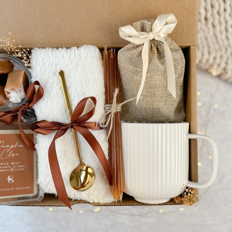 May include: A gift box containing a selection of comforting items. It features a white mug, a small hessian bag tied with a cream ribbon, a fluffy white blanket, a gold spoon, brown candles, and caramel sweets. A candle wax melt is also included.