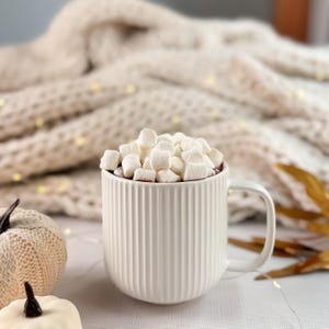 May include: A white ceramic mug filled with hot chocolate and topped with marshmallows. The mug has vertical ribbed detailing and a handle. Two small white pumpkins and a beige knitted blanket are in the background.