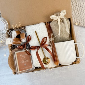 May include: A gift box containing a white mug, a small hessian bag tied with a cream ribbon, a white fluffy towel, a gold spoon, a candle, and caramel sweets. The candle is labelled "Pumpkin Clove". The box is brown.