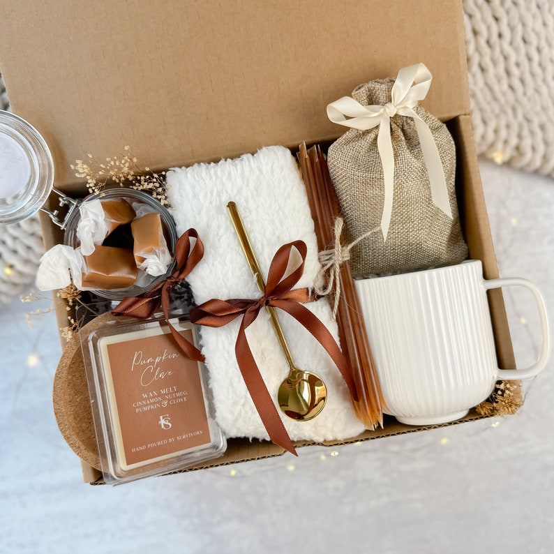 May include: A gift box containing a white mug, a small hessian bag tied with a cream ribbon, a white fluffy towel, a gold spoon, a candle, and caramel sweets. The candle is labelled "Pumpkin Clove". The box is brown.