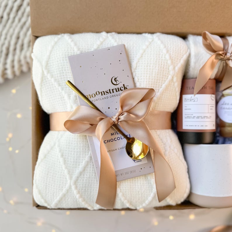May include: A gift box with a cream-colored waffle-knit blanket tied with a satin ribbon. A gold spoon rests on a chocolate bar with the text "moonstruck" and "MILK CHOCOLATE". Other items include a tea tin and a mug.