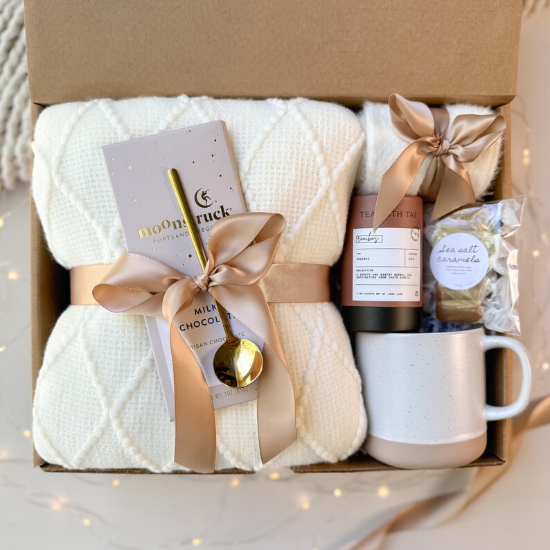 May include: A gift box filled with a cream-colored waffle-knit blanket, a chocolate bar with a gold spoon, a mug, tea, and sea salt caramels. The items are tied with a satin ribbon.