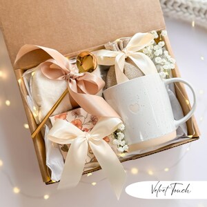 May include: A gift box with a white mug, a gold spoon, a small floral-patterned box, and a fluffy white item, all tied with peach-colored ribbons. The mug has a heart design and is filled with a beige base. The box is lined with white paper and contains the text "Velvet Touch".