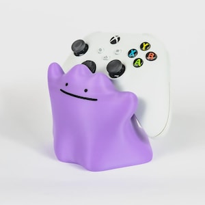 Könnte beinhalten: Ein viofter, klecksförmiger Controller-Ständer mit einem einfachen Gesicht aus zwei schwarzen Punkten als Augen und einem horizontalen Strich als Mund. Ein weißer kabelloser Game-Controller mit schwarzen Joysticks und bunten Tasten steht darin.
