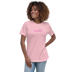 Może przedstawiać: Różowy t-shirt z napisem "Powered by Pilates" w czarnym i różowym kolorze.
