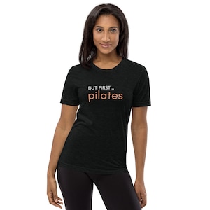 Może przedstawiać: Czarny T-shirt z napisem "BUT FIRST... pilates" w kolorze białym i pomarańczowym.