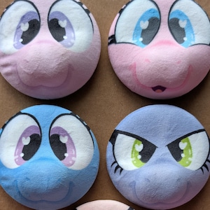May include: Five circular plush patches featuring cartoon faces with large expressive eyes. The top row includes a light pink patch with purple eyes and a pink patch with blue eyes. The middle row has a blue patch with purple eyes and a lavender patch with green eyes. A bottom pink patch has orange eyes.