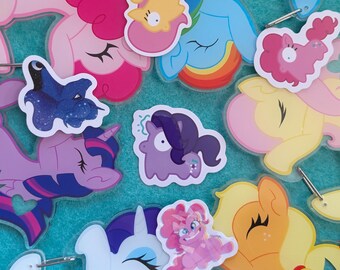 Pode incluir: Uma coleção de pingentes de acrílico com vários personagens de My Little Pony em diferentes poses e cores, incluindo pôneis rosa, azuis, amarelos e roxos, com alguns pingentes exibindo pequenos anéis de metal para fixação.