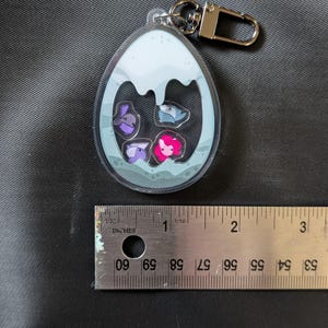 Puede incluir: Llavero acr&iacute;lico transparente con cierre plateado. El llavero presenta un dise&ntilde;o en forma de huevo con un fondo azul claro y cuatro personajes de dibujos animados en morado, azul y rosa. El llavero mide aproximadamente 5 cm de largo.
