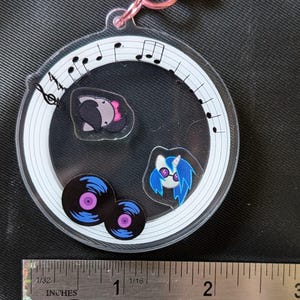 Puede incluir: Un llavero redondo y transparente con un cierre rosa. En el interior, hay im&aacute;genes de dibujos animados de un animal gris con un lazo rosa, un pony de pelo azul y dos discos de vinilo. El llavero est&aacute; decorado con notas musicales.