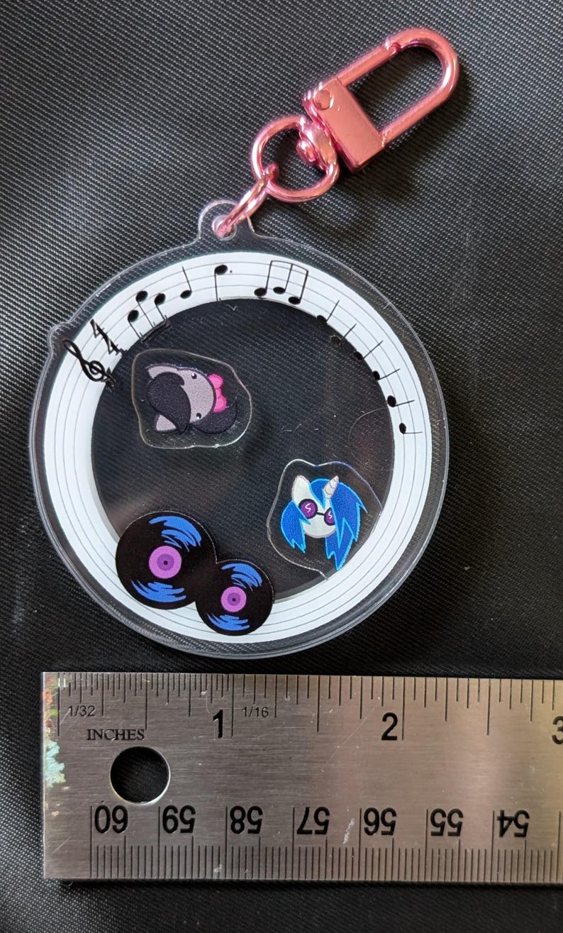 Puede incluir: Un llavero redondo y transparente con un cierre rosa. En el interior, hay im&aacute;genes de dibujos animados de un animal gris con un lazo rosa, un pony de pelo azul y dos discos de vinilo. El llavero est&aacute; decorado con notas musicales.