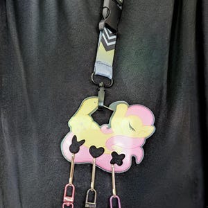 Pode incluir: Um cord&atilde;o com um desenho de p&ocirc;nei de desenho animado, com um pingente grande e pingentes menores. O cord&atilde;o tem um fecho preto e uma al&ccedil;a estampada. O pingente grande &eacute; amarelo e rosa, com tr&ecirc;s pingentes menores pendurados abaixo.