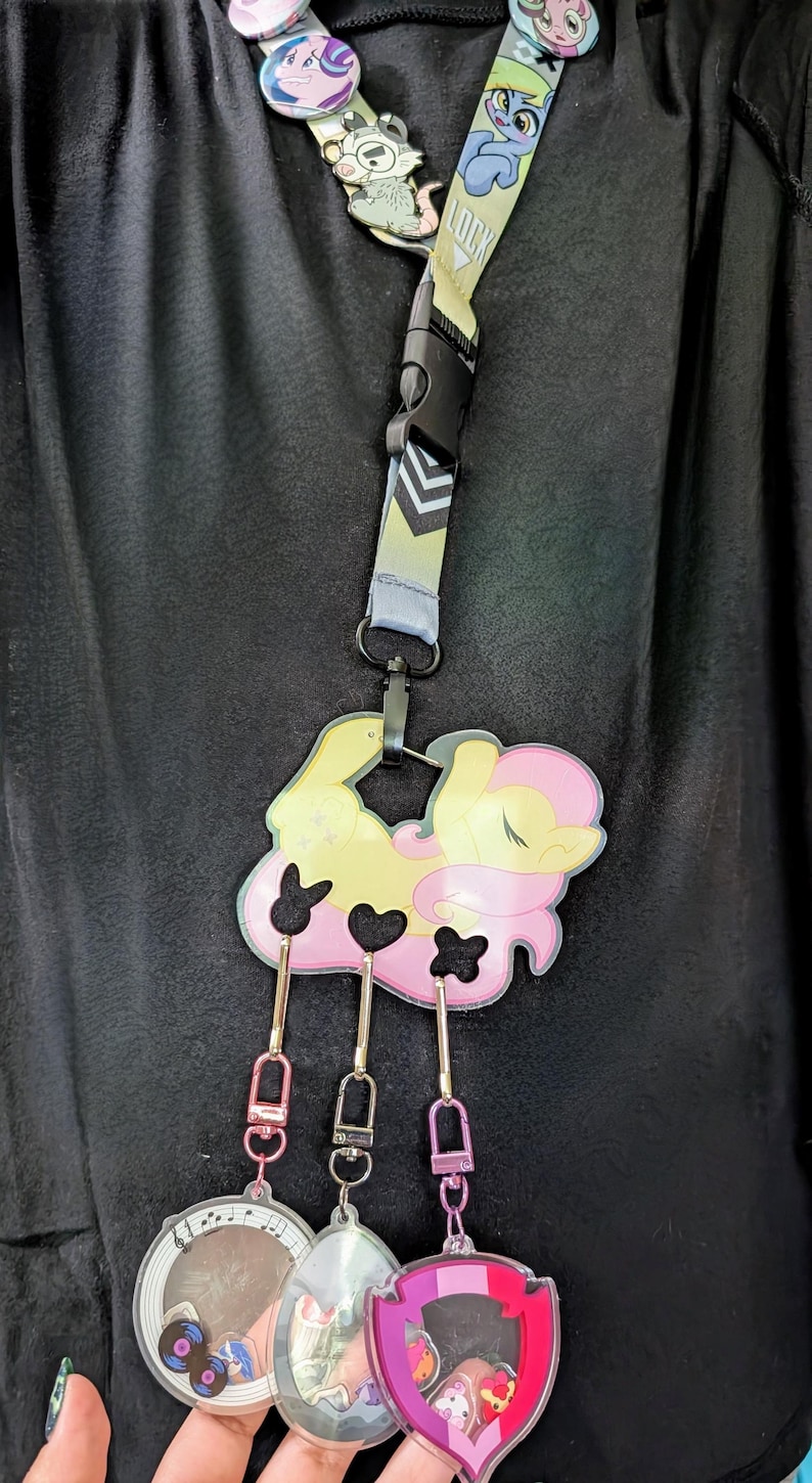 Pode incluir: Um cord&atilde;o com um desenho de p&ocirc;nei de desenho animado, com um pingente grande e pingentes menores. O cord&atilde;o tem um fecho preto e uma al&ccedil;a estampada. O pingente grande &eacute; amarelo e rosa, com tr&ecirc;s pingentes menores pendurados abaixo.