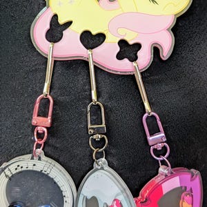 Pode incluir: Um porta-chaves com v&aacute;rios berloques com um personagem de desenho animado amarelo e rosa. O porta-chaves tem tr&ecirc;s berloques: um berloque com tema musical, um berloque cinza e um berloque rosa. Cada berloque tem um design diferente.