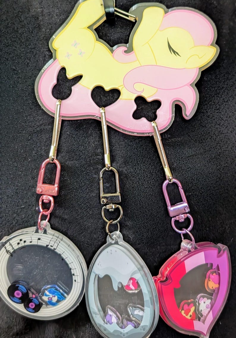 Pode incluir: Um porta-chaves com v&aacute;rios berloques com um personagem de desenho animado amarelo e rosa. O porta-chaves tem tr&ecirc;s berloques: um berloque com tema musical, um berloque cinza e um berloque rosa. Cada berloque tem um design diferente.