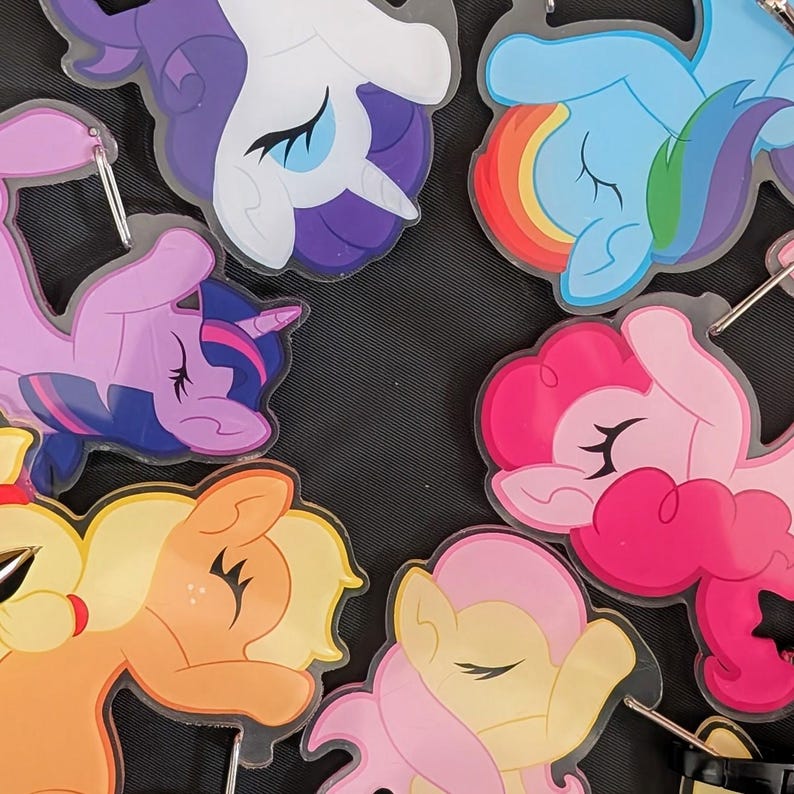 Pode incluir: Uma cole&ccedil;&atilde;o de chaveiros coloridos em forma de p&ocirc;nei de desenho animado. Os chaveiros apresentam v&aacute;rios p&ocirc;neis em tons de roxo, rosa, amarelo, azul e laranja. Cada chaveiro tem um anel de metal prateado.