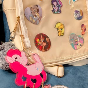 Pode incluir: Mochila creme com um painel frontal transparente exibindo v&aacute;rios pinos de personagens de desenhos animados. Um grande chaveiro rosa em forma de p&ocirc;nei est&aacute; preso, junto com outros chaveiros. A bolsa tem z&iacute;peres e ferragens dourados.