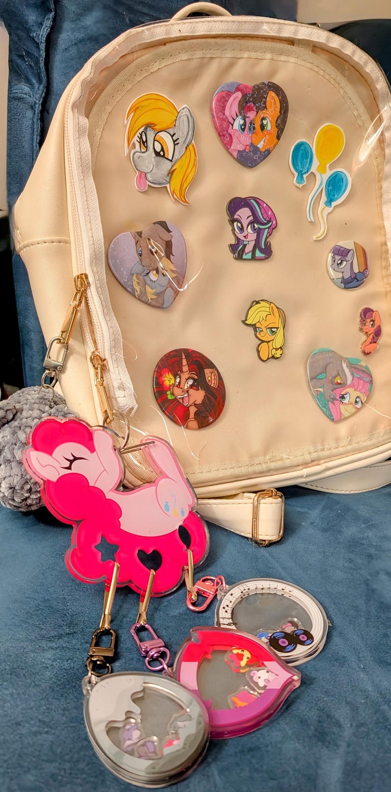 Pode incluir: Mochila creme com um painel frontal transparente exibindo v&aacute;rios pinos de personagens de desenhos animados. Um grande chaveiro rosa em forma de p&ocirc;nei est&aacute; preso, junto com outros chaveiros. A bolsa tem z&iacute;peres e ferragens dourados.