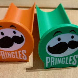 May include: Two Pringles-themed chip holders, one orange and one green, are shown. Each holder features the Pringles logo with a smiling face and mustache, and the word "PRINGLES" below. The holders are on a wooden surface.