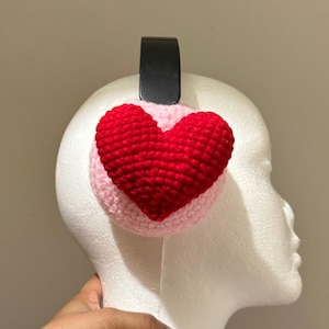 Op de afbeelding: Roze en rode gehaakte oorwarmers met een hartvorm. De oorwarmers zijn bevestigd aan een zwart hoofdband.