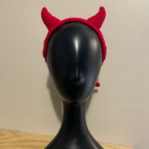 May include: Red crocheted headband with devil horns. The headband is on a black mannequin head.
