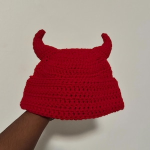 May include: A red crocheted hat with two curved horns. The hat is made of a thick, textured yarn and has a rounded crown and brim. The horns are also crocheted and are a darker shade of red.