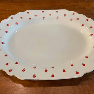 May include: An oval-shaped white serving platter with a scalloped edge and a repeating pattern of small red flowers with green stems scattered across the rim. The platter features a thin metallic silver trim along the outer scalloped border.