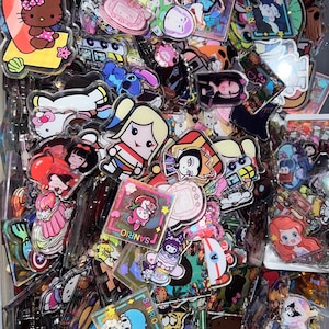 May include: A collection of colorful, clear acrylic charms featuring various cartoon characters, including Hello Kitty, a mermaid, and a superhero. The charms are decorated with glitter and other embellishments. The charms are perfect for adding a touch of whimsy to your keychain, bag, or phone.
