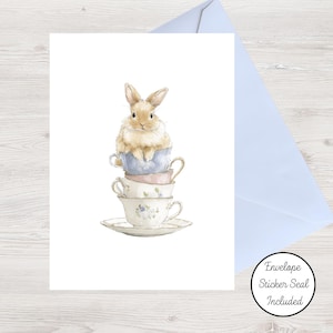 May include: A greeting card featuring a watercolor illustration of a light brown rabbit sitting inside a stack of three pastel-colored teacups on a saucer. A light blue envelope is positioned behind the card, and a circular sticker reads Envelope Sticker Seal Included.