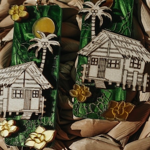 May include: Handmade earrings featuring a tropical theme. Each earring has a green background with a wooden cutout of a palm tree, a small house, and a yellow flower. The earrings are suspended from gold-colored hooks.