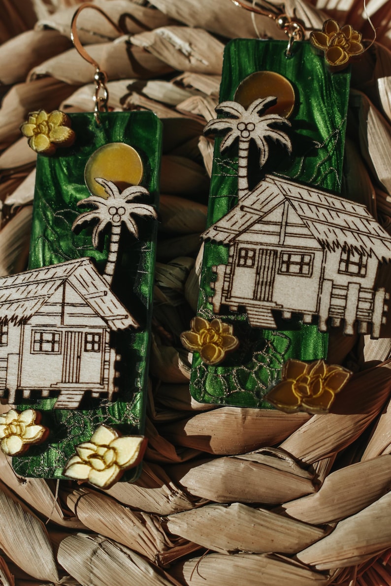 May include: Handmade earrings featuring a tropical theme. Each earring has a green background with a wooden cutout of a palm tree, a small house, and a yellow flower. The earrings are suspended from gold-colored hooks.