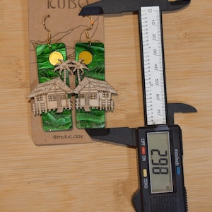 May include: Handmade earrings featuring a tropical design. The earrings have a green background with a gold sun and palm trees, and a wooden house. The earrings measure 2.98 inches in length.