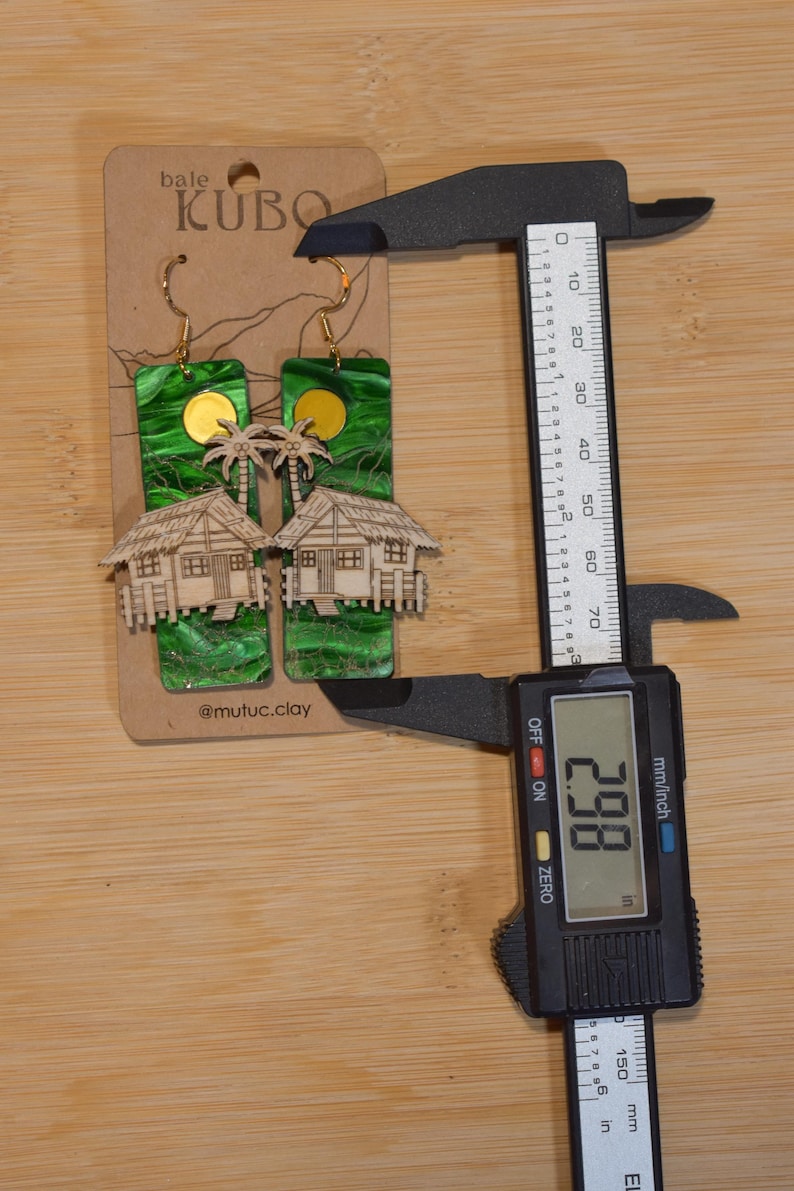 May include: Handmade earrings featuring a tropical design. The earrings have a green background with a gold sun and palm trees, and a wooden house. The earrings measure 2.98 inches in length.