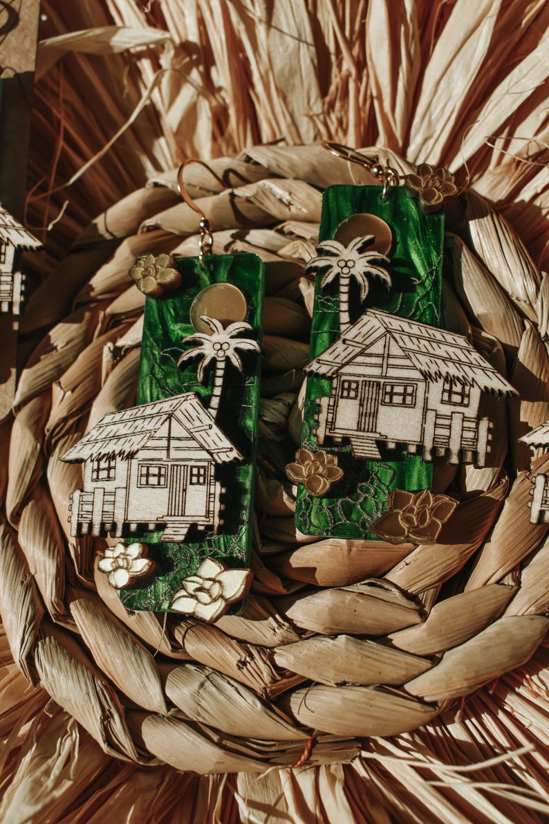 May include: Handmade earrings featuring a tropical theme. The earrings have a green background with a palm tree and a small house design. Gold-colored accents include a sun, flower, and earring hooks. The earrings are displayed on a woven, natural-colored surface.