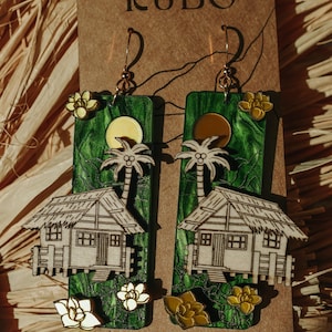 May include: Handmade wooden earrings featuring a tropical scene with a house, palm tree, and sun. The earrings have a green background with white and yellow accents. The text "bale KUBO" is at the top.