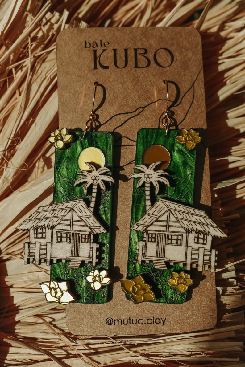 May include: Handmade wooden earrings featuring a tropical scene with a house, palm tree, and sun. The earrings have a green background with white and yellow accents. The text "bale KUBO" is at the top.