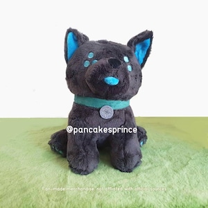 Puede incluir: Un peluche de perro en tonos grises y turquesas. El perro tiene orejas, nariz y detalles turquesas alrededor de los ojos. Lleva un collar turquesa con un colgante plateado. El texto "@pancakesprince" es visible.