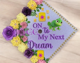 May include: A decorated graduation cap with a light purple glitter background. The cap features the text "On to My Next Dream" and a green chameleon. It is adorned with yellow, purple, and pink paper flowers and gold accents.