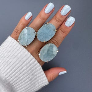 May include: Three large oval rings with pale blue gemstones are displayed on a hand with light blue nail polish. The rings have silver and gold accents. The hand is wearing a white sweater, set against a grey background.