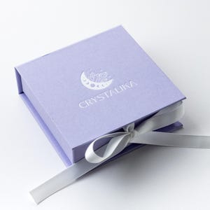 May include: A lavender gift box with a white ribbon and the logo "CRYSTALIKA" embossed on the top. The logo features a crescent moon with crystals inside.