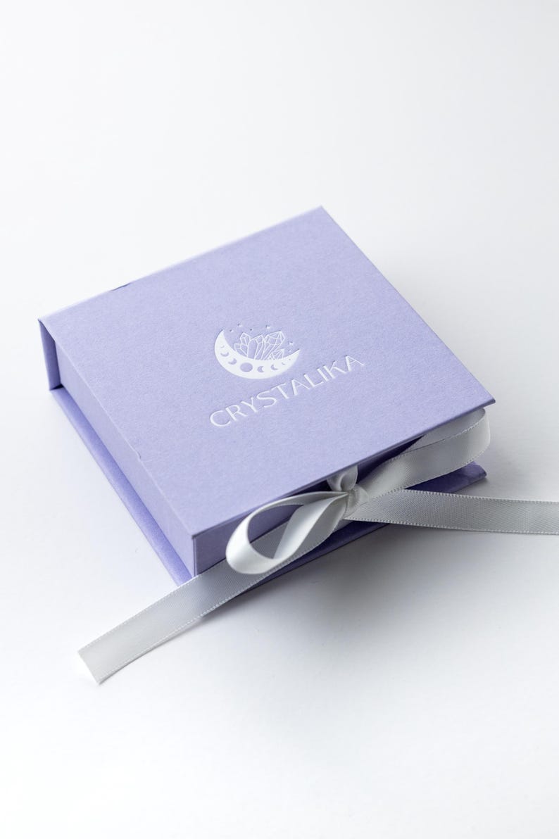 May include: A lavender gift box with a white ribbon and the logo "CRYSTALIKA" embossed on the top. The logo features a crescent moon with crystals inside.