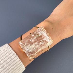 May include: A gold-toned metal cuff bracelet featuring a large, rectangular, clear crystal. The crystal has a textured, iridescent appearance. The bracelet is worn on a wrist, with a gray background.