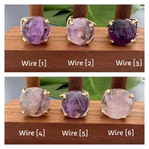May include: Six amethyst rings with gold wire settings displayed on a wooden stand. The rings are arranged in two rows of three. The amethyst stones are various shades of purple and pink.