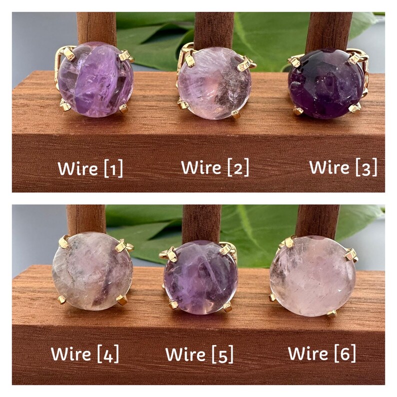 May include: Six amethyst rings with gold wire settings displayed on a wooden stand. The rings are arranged in two rows of three. The amethyst stones are various shades of purple and pink.