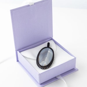 May include: An oval pendant with a gray and white stone is displayed inside a lavender gift box. The pendant has a black woven border and a black cord. The box is open, revealing a white satin lining and a white ribbon.